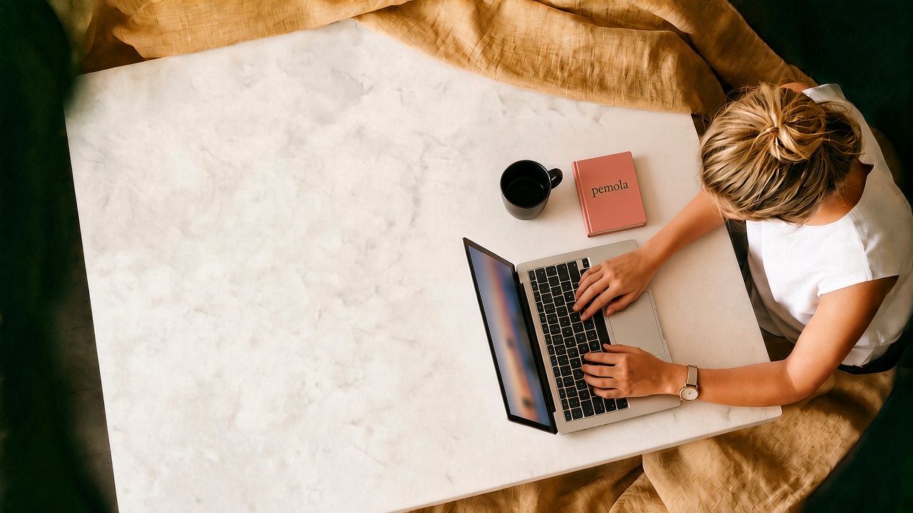Overhead view of a person working on a laptop at a marble table, a coffee and a pemola notebook beside them